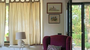 Ein Zimmer mit einem violetten Sessel, einem kleinen Tisch mit einer Lampe, einem Stuhl, einem Fenster mit Vorhängen und zwei gerahmten Bildern an der Wand.