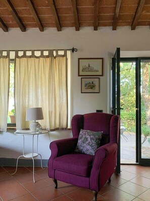 Ein Zimmer mit einem violetten Sessel, einem kleinen Tisch mit einer Lampe, einem Stuhl, einem Fenster mit Vorhängen und zwei gerahmten Bildern an der Wand.