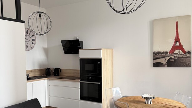 Uma cozinha moderna com forno embutido, uma área de refeições com mesa e cadeiras de madeira, e uma sala de estar com sofá.