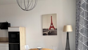 Uma cozinha moderna com micro-ondas, forno e armário de madeira. Uma mesa de jantar com cadeiras está centralizada no espaço, com uma luminária e uma obra de arte da Torre Eiffel na parede.