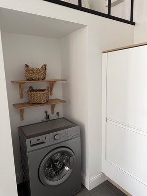 Un espace buanderie comprenant un lave-linge, deux étagères en bois avec des paniers et un mur blanc.