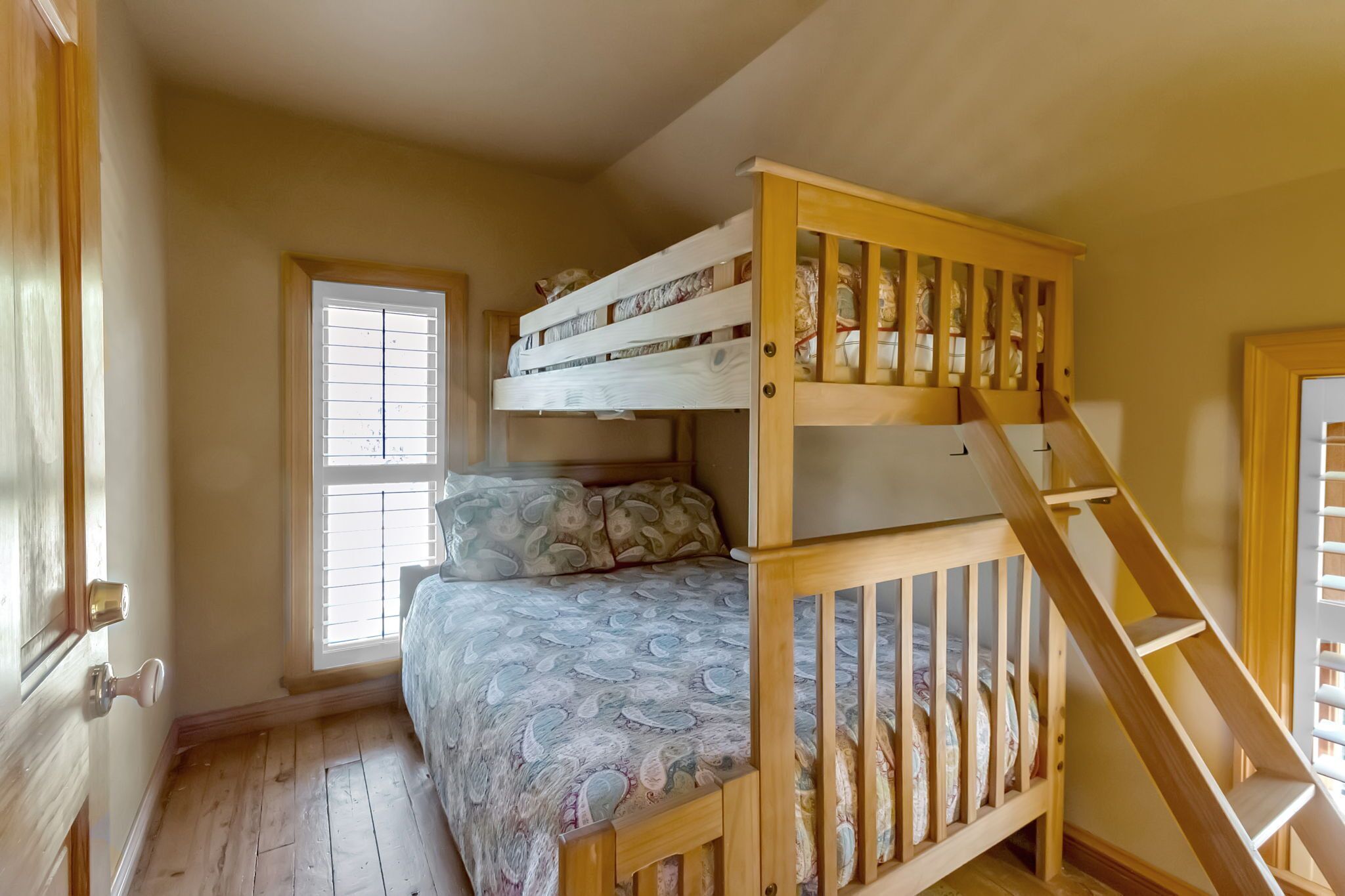 Une chambre avec un lit superposé, un escalier en bois, une fenêtre avec des stores et une porte.