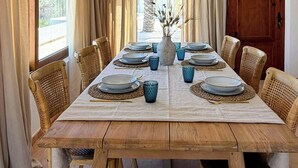 A dining room with a wooden table set for six, surrounded by wicker chairs, a hanging light fixture, and a view through a window with curtains. - Mediterranean-style house in Cabo de Gata (Almeria)