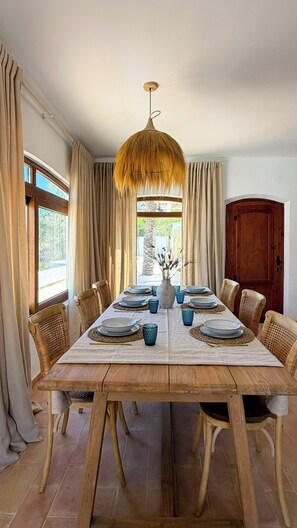 Ein Esszimmer mit einem Holztisch, gedeckt für sechs Personen, umgeben von Korbstühlen, einer Hängelampe und Blick durch ein mit Vorhängen drapiertes Fenster.