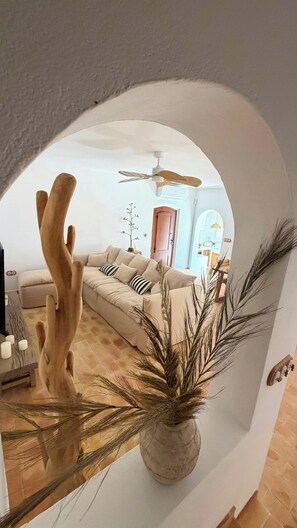 Ein bogenförmiger Durchgang führt in ein Wohnzimmer mit Sofa, Deckenventilator und einer Vase mit Trockenpflanzen.