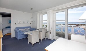 Ein Zimmer mit einem blauen Sofa, Korbstühlen, einem weißen Tisch und Meerblick durch große Fenster.