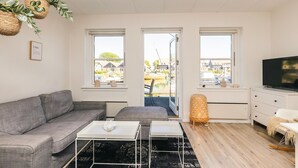 A modern living room with a grey sofa, white coffee table, and a TV on a dresser. There are three windows with views of a dock and boats. - Mon - Klintholm Harbour (Borre)