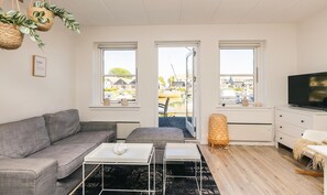 Ein modernes Wohnzimmer mit grauem Sofa, weißem Couchtisch und einem Fernseher auf einer Kommode. Drei Fenster bieten Blick auf einen Steg und Boote.