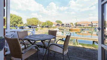 Une terrasse en bois, agrémentée de chaises et d’une table en osier, qui offre une vue sur une marina avec des bateaux et des bâtiments.