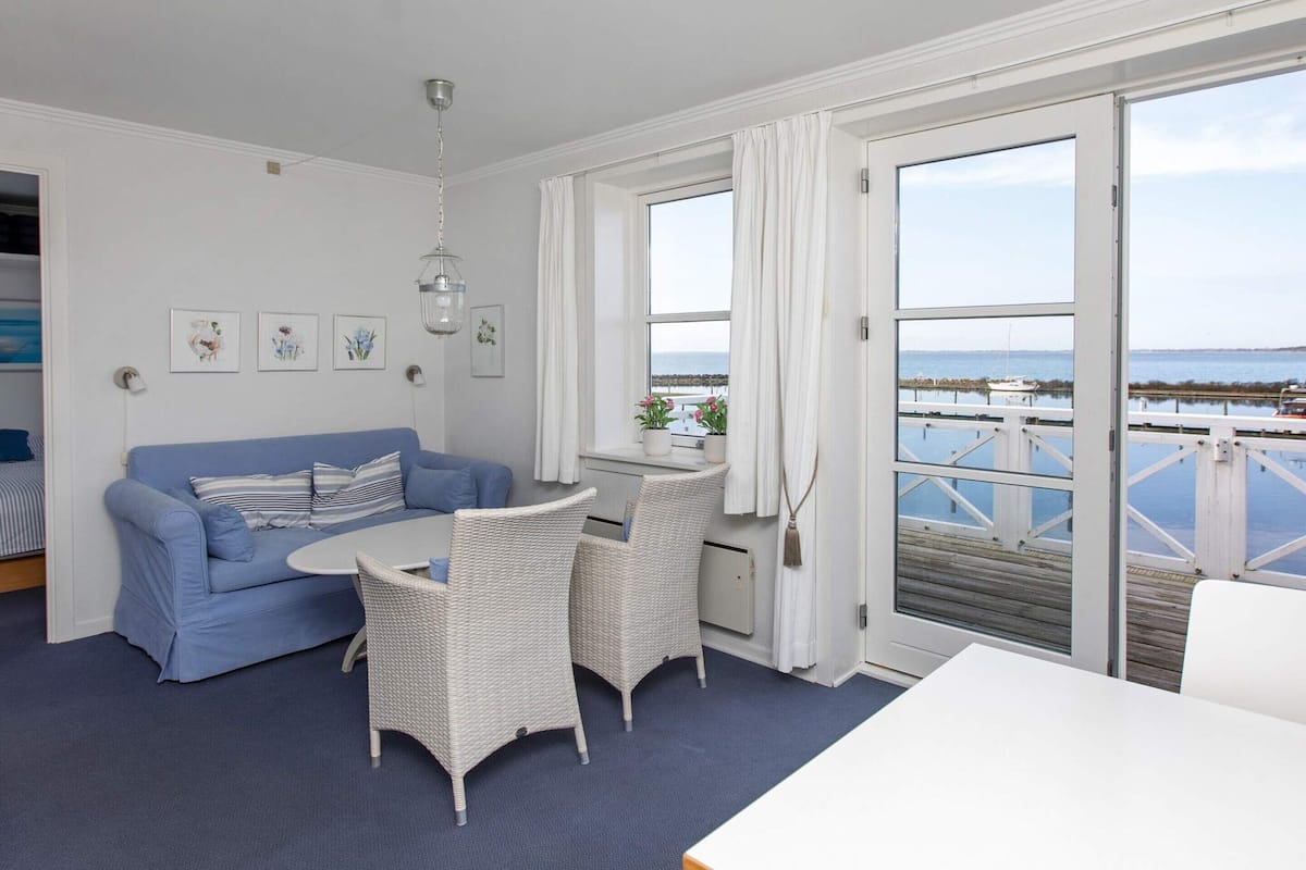 A room with a blue sofa, white wicker chairs, a small table, and a view of the sea through large windows.