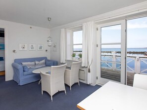 Ein Zimmer mit einem blauen Sofa, weißen Korbstühlen, einem kleinen Tisch und Meerblick durch große Fenster.