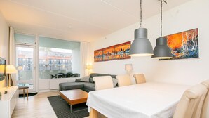 Ett modernt hotellrum med en säng, ett matbord, en soffa och ett stort fönster med utsikt över byggnader.