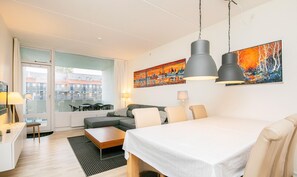 A modern hotel room with a bed, a dining table, a sofa, and a large window with a view of buildings.