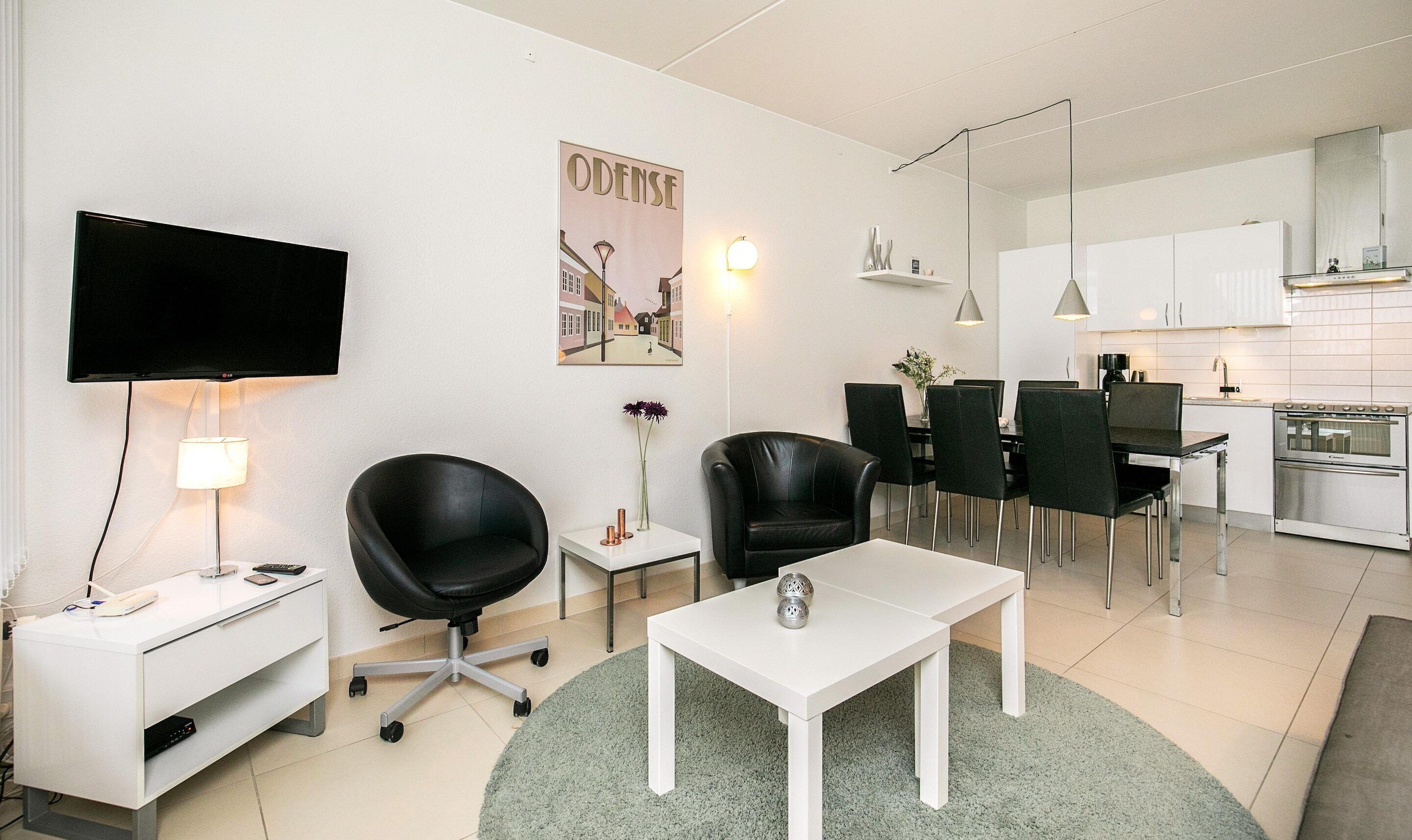 A modern living room with a flat-screen TV, a coffee table, a black leather chair, a dining area with chairs, and a kitchen with a stove.