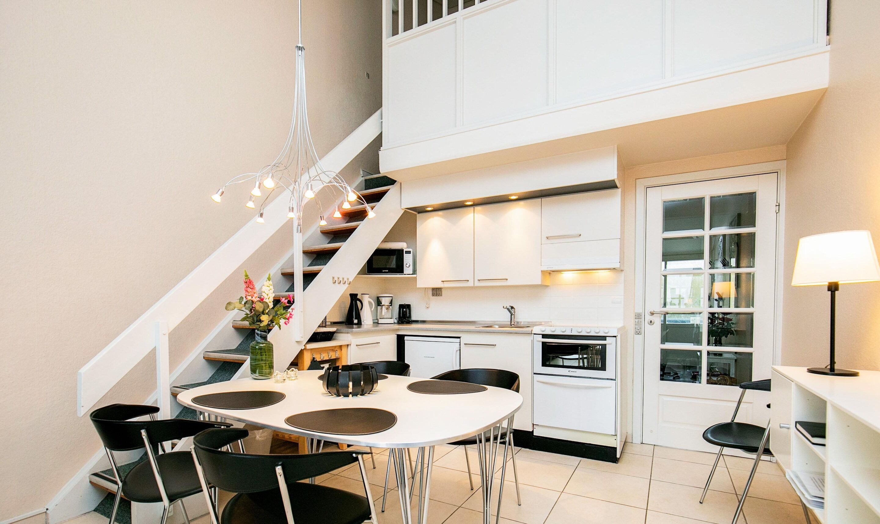Una cocina moderna con armarios blancos, un comedor con una mesa redonda y sillas, y una escalera que sube a un nivel superior.