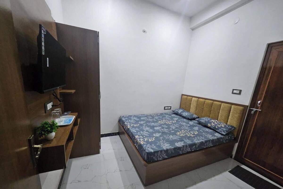 Un dormitorio con cama, televisor montado en la pared, mesita de noche con planta y una puerta.