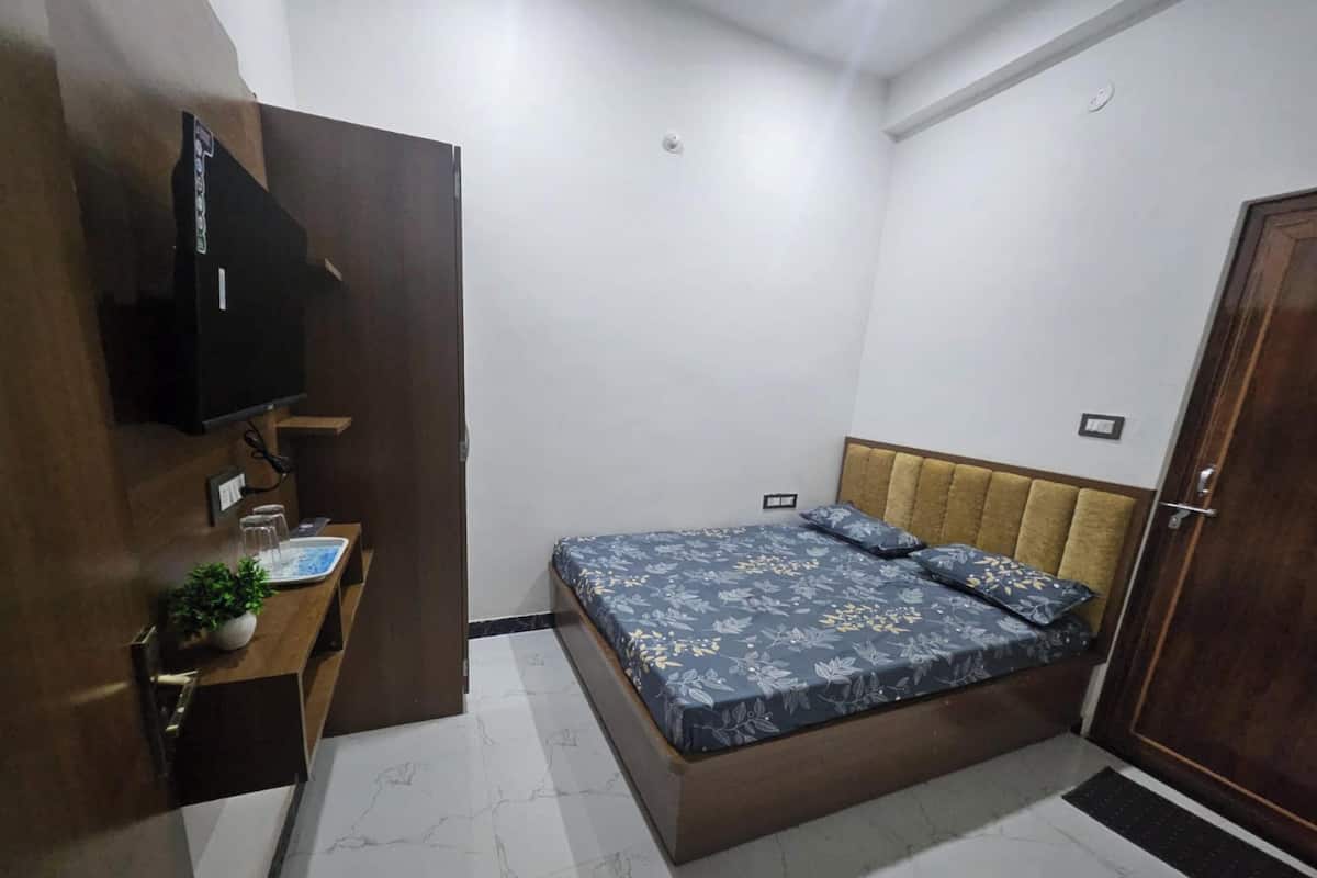 Quarto com cama, televisão fixada na parede, mesa de cabeceira com planta e porta.