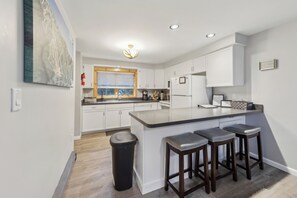Eine moderne Küche mit weißen Schränken, einer großen Kochinsel mit Barhocker, einem Fenster mit Jalousien und einer Deckenleuchte.