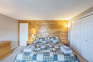 Ein Schlafzimmer mit Holzkopfende, einem Bett mit kariertem Überwurf, einem Nachttisch mit Lampe, einer Kommode und einem Schrank mit weißen Türen.
