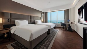 Quarto de hotel com duas camas, uma escrivaninha, uma cadeira, uma televisão e uma grande janela com vista para a cidade.