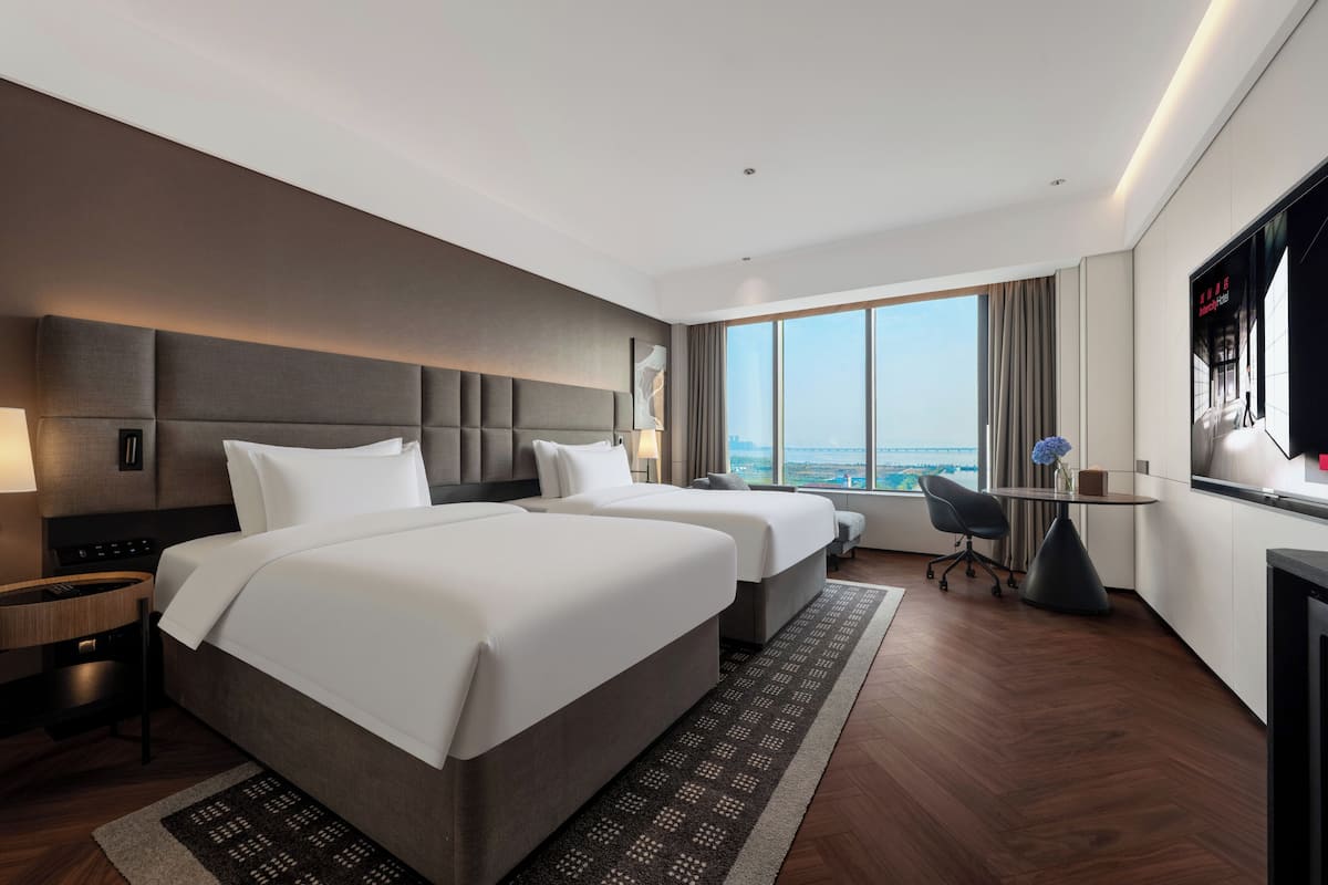 A hotel room with two beds, a desk, a chair, a TV, and a large window with a city view.