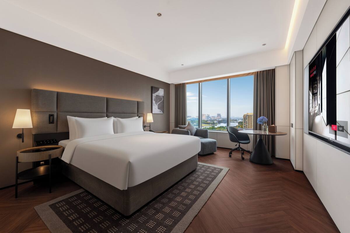 A modern hotel room with a large bed, a desk, a chair, a view of the city, and a large window.