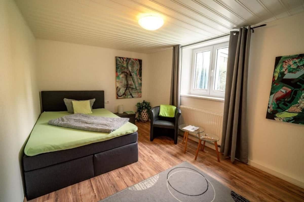 A bedroom with a bed, a chair, a small table, a radiator, and a window with curtains.