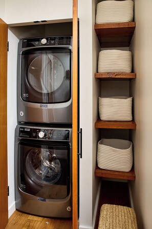 Kompakti pyykinhuoltotila, jossa on etukuormitteinen Maytag-pesukone ja -kuivausrumpu, puiset hyllyt säilytyskoreineen sekä puulattia.
