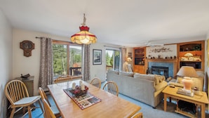 A cozy living room with a fireplace, dining area, and large windows. - Townhome Near Gore Mtn | Covered Heated Pool + Rec Lounge | Quick Silver (North Creek)