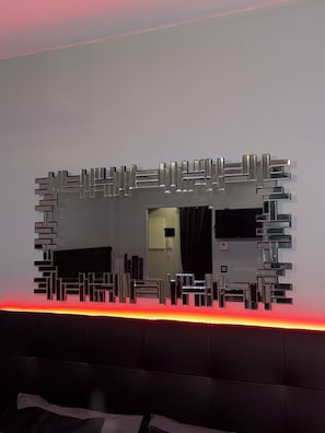 A modern room with a unique geometric mirror design on the wall, a dark upholstered headboard, and a red underlighting strip. - The sweet parenthesis (Dijon)