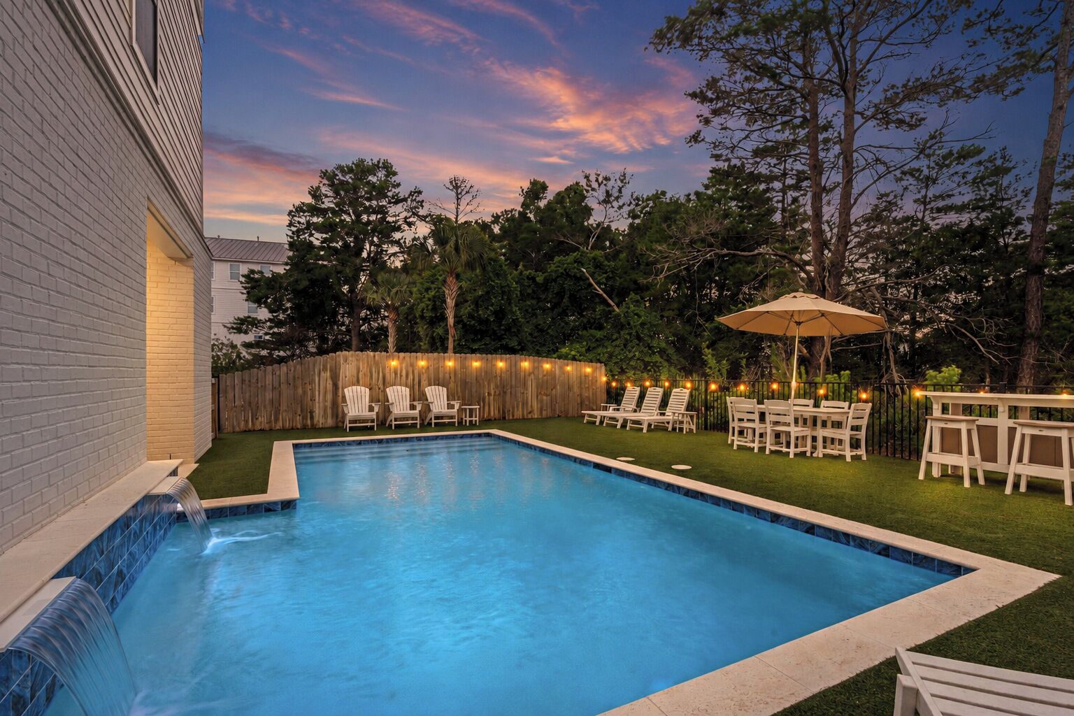 Una piscina con caída de agua, rodeada por una valla de madera y muebles de exterior.