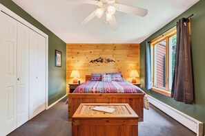 Een slaapkamer met een houten hoofdeinde, een bed met beddengoed met een patroon, twee bedlampen, een plafondventilator, een raam met gordijnen en een deur.