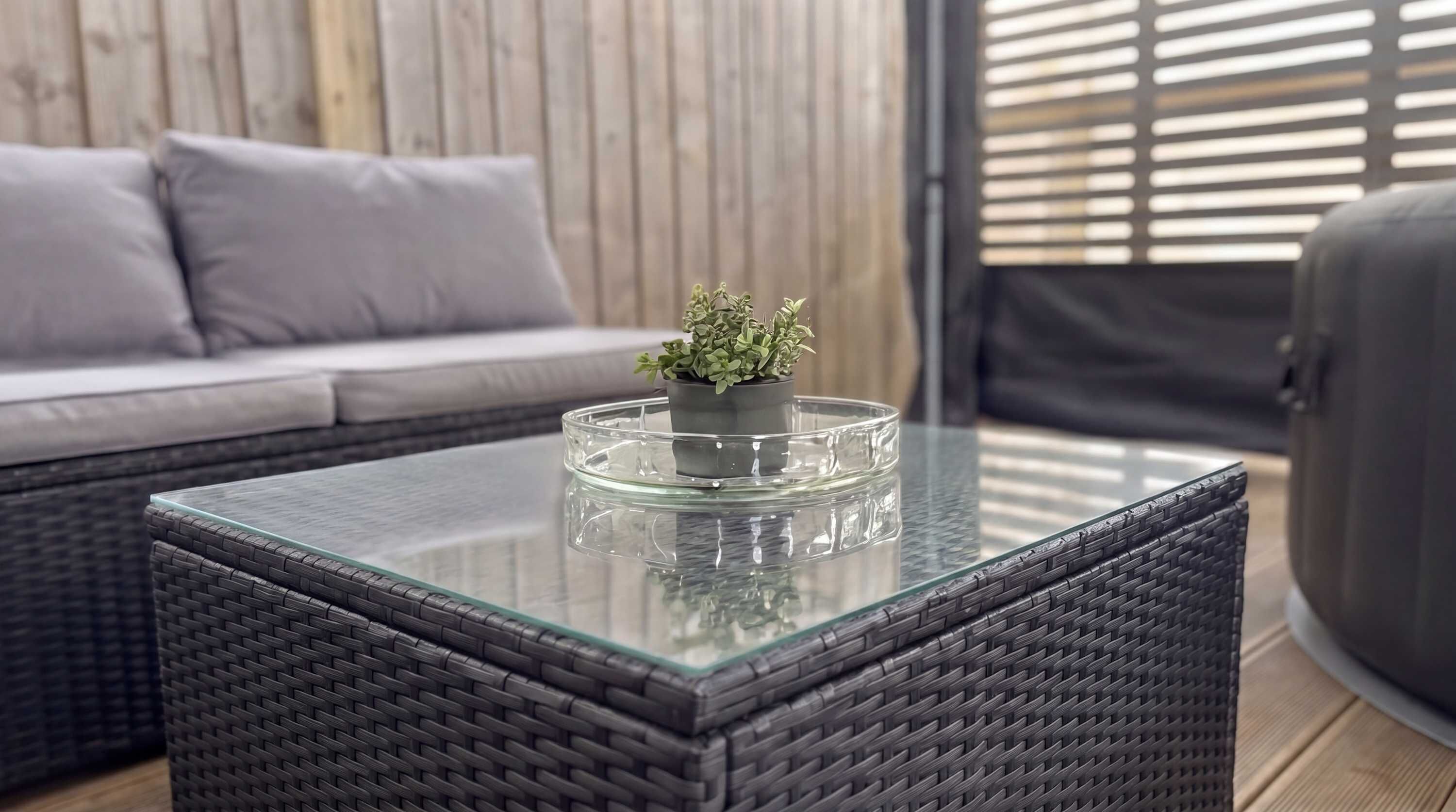 Uma mesa com tampo de vidro, um vaso com planta, um sofá de vime e uma cadeira de vime em um quarto com parede de madeira e persianas.