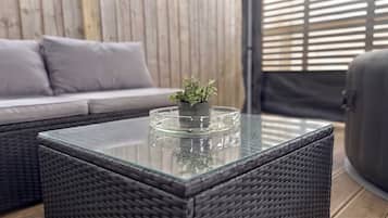 Uma mesa com tampo de vidro, um vaso com planta, um sofá de vime e uma cadeira de vime em um quarto com parede de madeira e persianas.