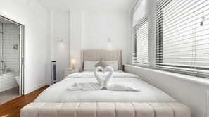 A hotel room with a bed, a bedside table, a nightstand, a window with blinds, and a bathroom visible through an open door. - VOLCANIC HOT TUB EXPERIENCE Central London 2BR Lagoon, Memories Await for 6 (Greater London)