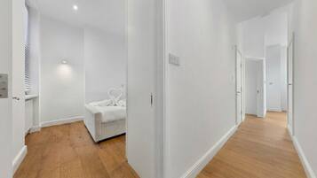 Um corredor com piso de madeira, paredes brancas e uma cama com roupa de cama branca.