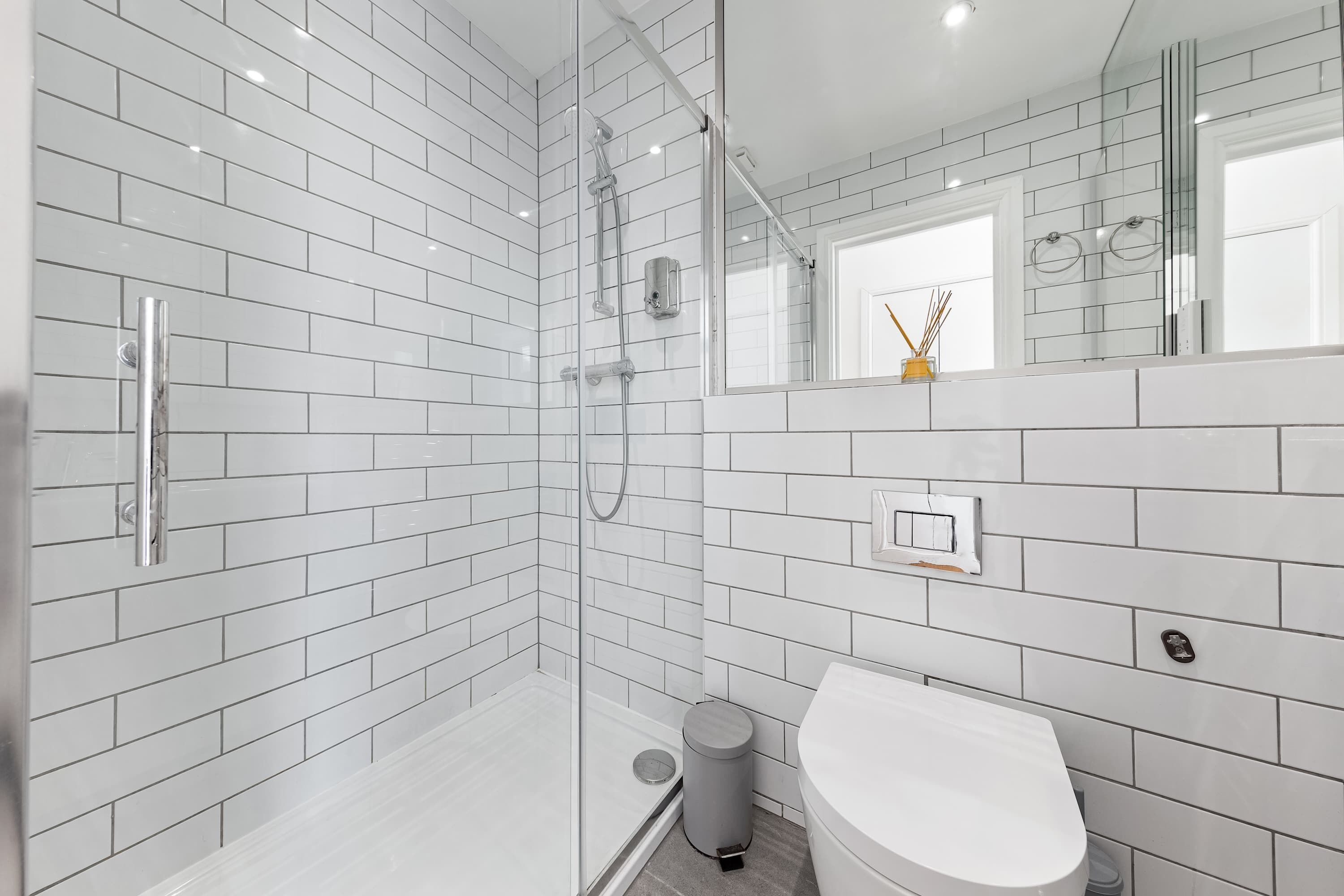 Une salle de bain moderne avec des carreaux de céramique blancs rectangulaires, une douche en verre, un lavabo blanc et un miroir agrémenté d’un petit objet décoratif fixé au mur.
