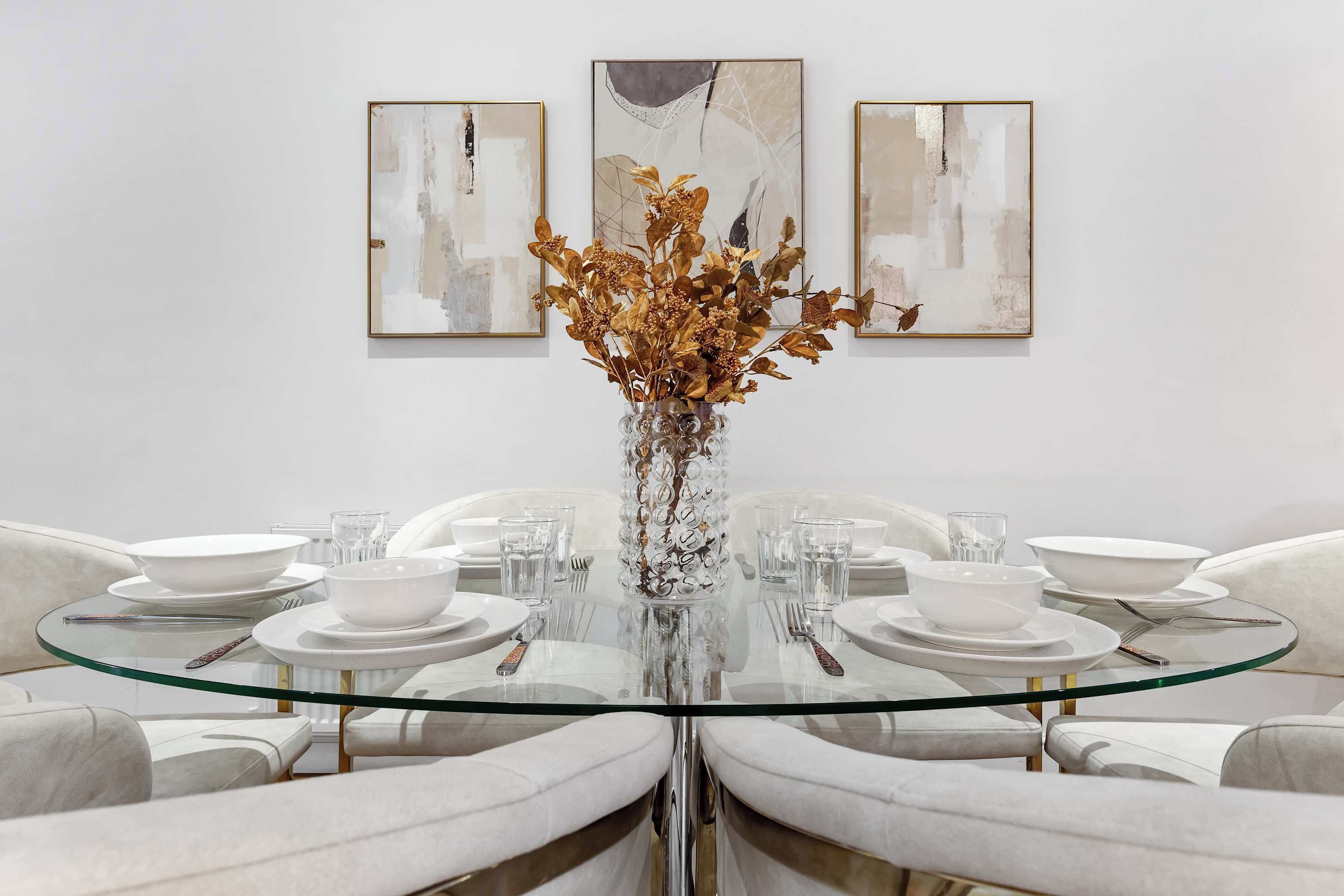 A dining table with a glass top surrounded by white chairs, set with white plates, bowls, and cutlery.