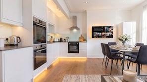 A modern kitchen with built-in appliances, a dining area with a table set for two, and a wall-mounted TV displaying a movie. - Shard View on Terrace 4BR Rare London Station Stay, Close to London Eye, 2 Min walk From Station, Cinematic Scenes for 10 (Greater London)