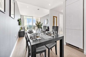 A modern dining area with a long table set for four, a large window, and a view of the outdoors. - Sky Scraper London Luxury 1BR Roof Terrace and Gym (Greater London)
