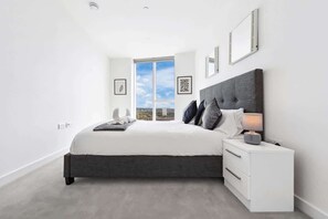 A modern bedroom with a large bed, a nightstand, a window with a view, and framed artwork on the wall. - Sky Scraper London Luxury 1BR Roof Terrace and Gym (Greater London)