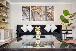 Un salon moderne avec un canapé noir, des coussins blancs, une table basse en verre et une œuvre d’art abstraite accrochée au mur.