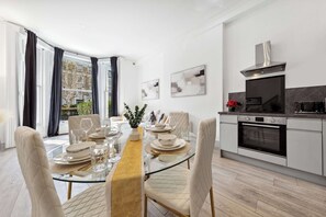 A modern dining room with a glass table, white chairs, and a view of the outdoors through large windows. - VIRAL Kensignton 2BR Refreshing Dream Home by Station for 6 (Greater London)