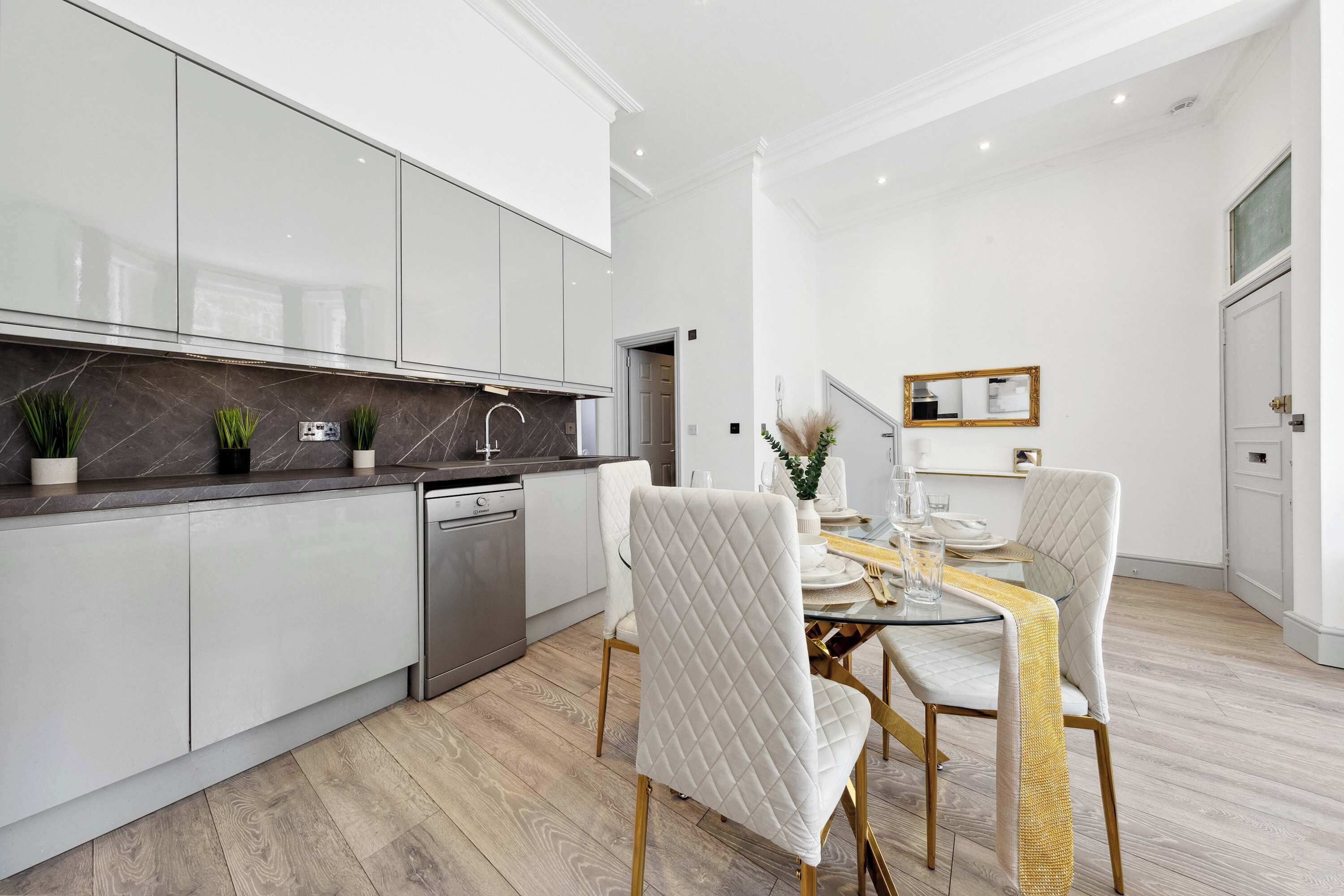 A modern kitchen with white cabinets, a built-in dishwasher, and a dining area with a glass table and white upholstered chairs.