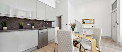 Une cuisine moderne avec des meubles blancs, un lave-vaisselle encastré et un coin repas comprenant une table en verre et des chaises à revêtement blanc.