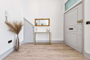 A hallway with a white door, a mirror on the wall, a small table with a picture frame, and a vase with dried plants. - VIRAL Kensignton 2BR Refreshing Dream Home by Station for 6 (Greater London)