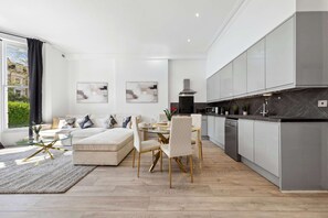 A modern living room with a sofa, dining table, and chairs. There is a kitchen with cabinets and a dishwasher. - VIRAL Kensignton 2BR Refreshing Dream Home by Station for 6 (Greater London)