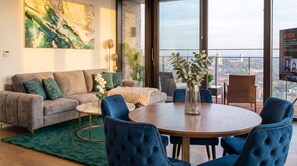 Ein modernes Wohnzimmer mit grauem Sofa, blauen Sesseln, einem runden Holztisch und einem großen Fenster mit Blick auf die Stadt.