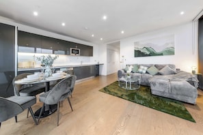 A modern living room with a grey sofa, a dining area with a table set for four, and a kitchen with dark cabinets and a microwave. - Tik Tok Famous Skyline Apartment 2BR Memories London, One of London's Most Exclusive Stays (Greater London)