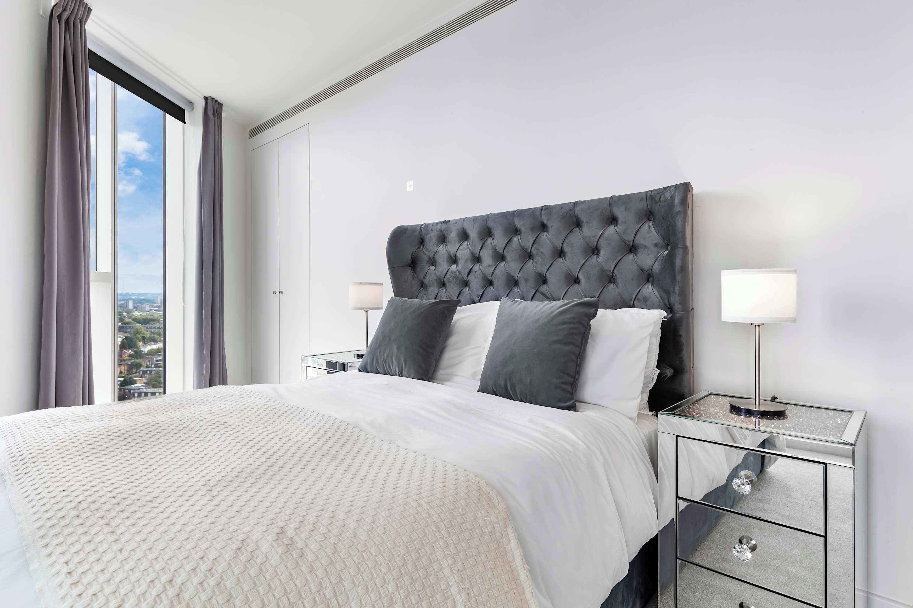 Habitación de hotel moderna con una cama grande, cabecera gris y vista a la ciudad por la ventana.